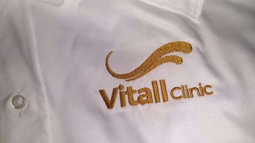Zbliżenie złotego haftu logo Vitall Clinic na białej koszuli